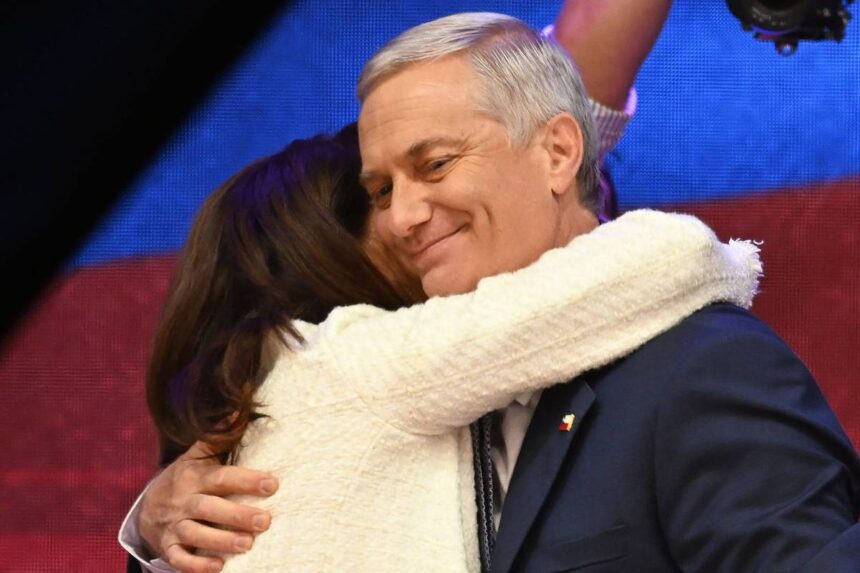 Chile: Kast é eleito e direita retorna ao poder em 2025 Homem de cabelos grisalhos veste terno azul escuro e sorri com os olhos fechados enquanto é abraçado por mulher de cabelos castanhos e casaco branco. Fundo desfocado com luzes coloridas.