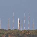 Foguete branco posicionado verticalmente em plataforma de lançamento no topo de área com vegetação densa. Antenas altas com faixas vermelhas e brancas cercam o foguete contra céu azul claro.