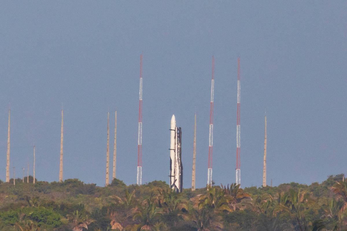 Foguete branco posicionado verticalmente em plataforma de lançamento no topo de área com vegetação densa. Antenas altas com faixas vermelhas e brancas cercam o foguete contra céu azul claro.