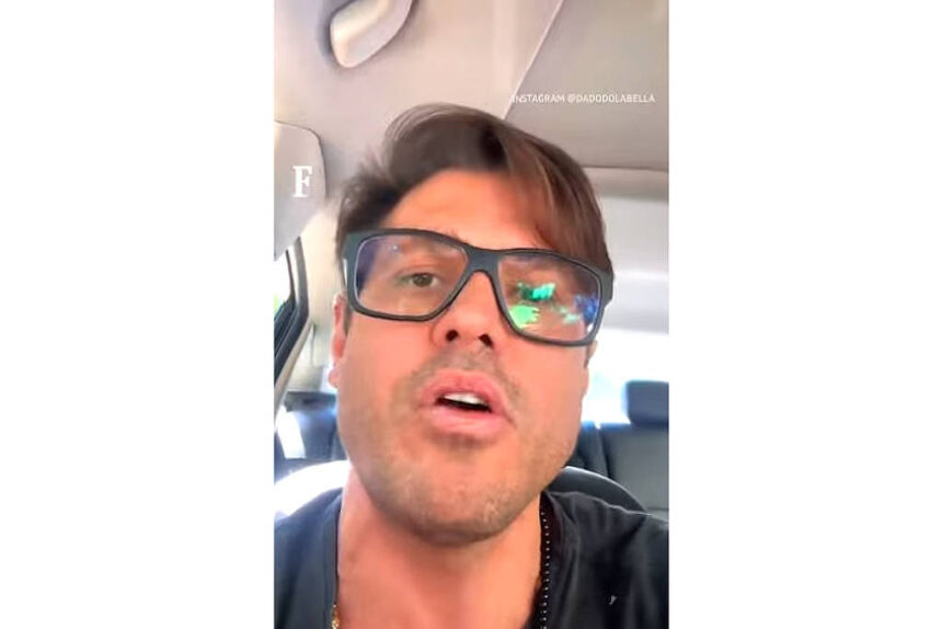 Homem com cabelo castanho claro e óculos escuros grandes está sentado no banco traseiro de um carro. Ele veste camiseta preta e o interior do veículo é claro, com bancos escuros ao fundo.