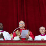 Papa ao centro com vestes vermelhas e douradas, segurando papéis e falando ao microfone; ao lado dele, dois religiosos de batina branca com gola roxa, diante de cortina vermelha.