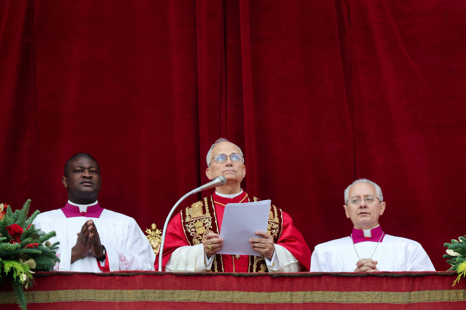 Papa ao centro com vestes vermelhas e douradas, segurando papéis e falando ao microfone; ao lado dele, dois religiosos de batina branca com gola roxa, diante de cortina vermelha.
