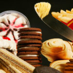 A imagem apresenta uma variedade de doces e sobremesas dispostos em uma mesa. Há uma taça de sobremesa com creme e cerejas, bolos com cobertura, biscoitos recheados, rolinhos de massa e batatas fritas em uma embalagem. Os doces estão organizados de forma a mostrar diferentes texturas e formas, com um fundo escuro que destaca os itens.