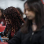 A imagem mostra um grupo de estudantes em uma sala de aula. Em primeiro plano, uma estudante com cabelo cacheado e vermelho está escrevendo em um caderno, enquanto outras pessoas estão visíveis ao fundo, algumas com expressões de concentração. O ambiente parece ser um espaço educacional, com cadeiras vermelhas e um fundo desfocado.