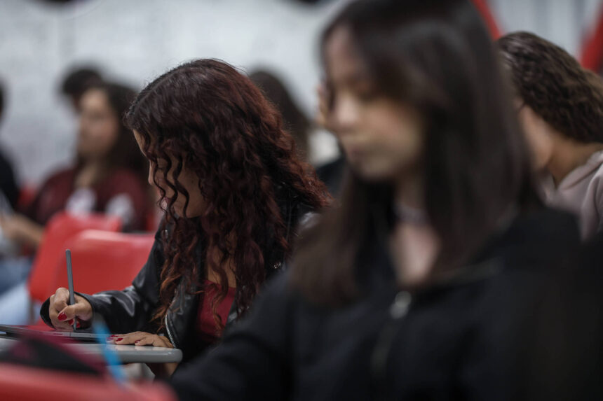 A imagem mostra um grupo de estudantes em uma sala de aula. Em primeiro plano, uma estudante com cabelo cacheado e vermelho está escrevendo em um caderno, enquanto outras pessoas estão visíveis ao fundo, algumas com expressões de concentração. O ambiente parece ser um espaço educacional, com cadeiras vermelhas e um fundo desfocado.