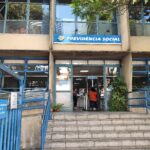 A imagem mostra a entrada de uma agência da Previdência Social. A fachada é de um prédio de dois andares, com janelas grandes e uma escada de acesso. Acima da entrada, há uma placa azul com o texto