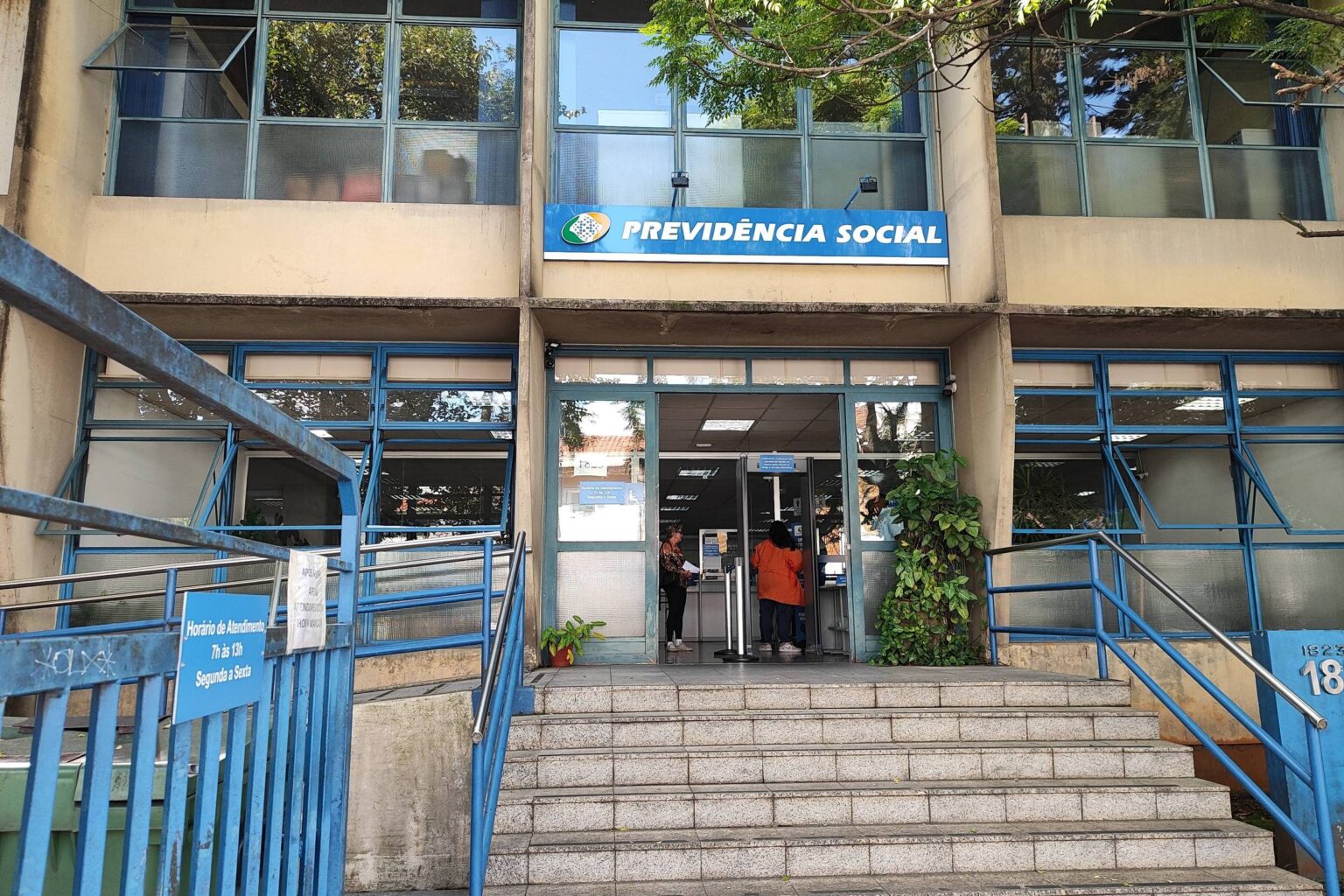 A imagem mostra a entrada de uma agência da Previdência Social. A fachada é de um prédio de dois andares, com janelas grandes e uma escada de acesso. Acima da entrada, há uma placa azul com o texto