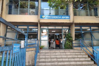 A imagem mostra a entrada de uma agência da Previdência Social. A fachada é de um prédio de dois andares, com janelas grandes e uma escada de acesso. Acima da entrada, há uma placa azul com o texto