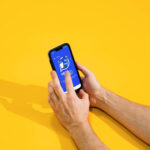 A imagem mostra duas mãos segurando um smartphone com a tela voltada para cima. O smartphone exibe um aplicativo com um fundo azul e um ícone central. O fundo da imagem é amarelo, criando um contraste vibrante.