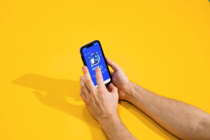 Calendário Bolsa Família tem início em 19 de janeiro A imagem mostra duas mãos segurando um smartphone com a tela voltada para cima. O smartphone exibe um aplicativo com um fundo azul e um ícone central. O fundo da imagem é amarelo, criando um contraste vibrante.