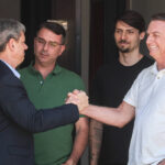 Quatro homens estão reunidos em ambiente interno. Dois deles, um de terno escuro e outro de camisa branca, apertam as mãos com expressão de cordialidade. Os outros dois, um de camisa verde e outro de camiseta preta, observam a interação. Fundo neutro com pouca iluminação.