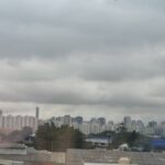 Vista panorâmica da cidade com prédios residenciais e comerciais sob um céu carregado de nuvens cinzentas. Áreas verdes e construções de diferentes alturas compõem a paisagem urbana.