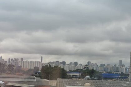Vista panorâmica da cidade com prédios residenciais e comerciais sob um céu carregado de nuvens cinzentas. Áreas verdes e construções de diferentes alturas compõem a paisagem urbana.