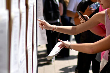 Sisu 2026: notas de corte da seleção anterior Braço de pessoa aponta para documento em painel com várias folhas afixadas. Outra pessoa ao fundo segura celular, ambiente externo com luz natural.
