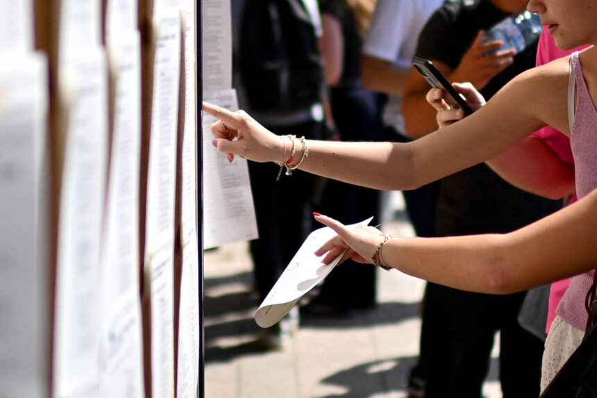 Braço de pessoa aponta para documento em painel com várias folhas afixadas. Outra pessoa ao fundo segura celular, ambiente externo com luz natural.