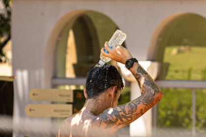 Homem sem camisa com tatuagens no braço e relógio no pulso despeja água de uma garrafa plástica na cabeça. Ao fundo, estrutura com arcos e placas de sinalização indicam ambiente externo.