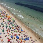 Vista aérea mostra praia cheia de guarda-sóis coloridos distribuídos na areia e muitas pessoas na água e na faixa de areia. O mar apresenta áreas claras e escuras, com ondas suaves chegando à praia.