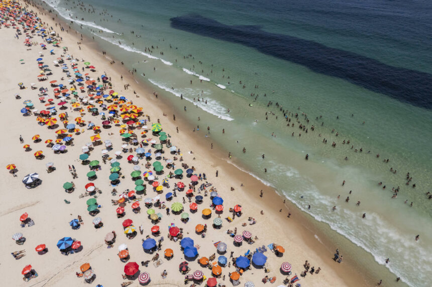 Vista aérea mostra praia cheia de guarda-sóis coloridos distribuídos na areia e muitas pessoas na água e na faixa de areia. O mar apresenta áreas claras e escuras, com ondas suaves chegando à praia.