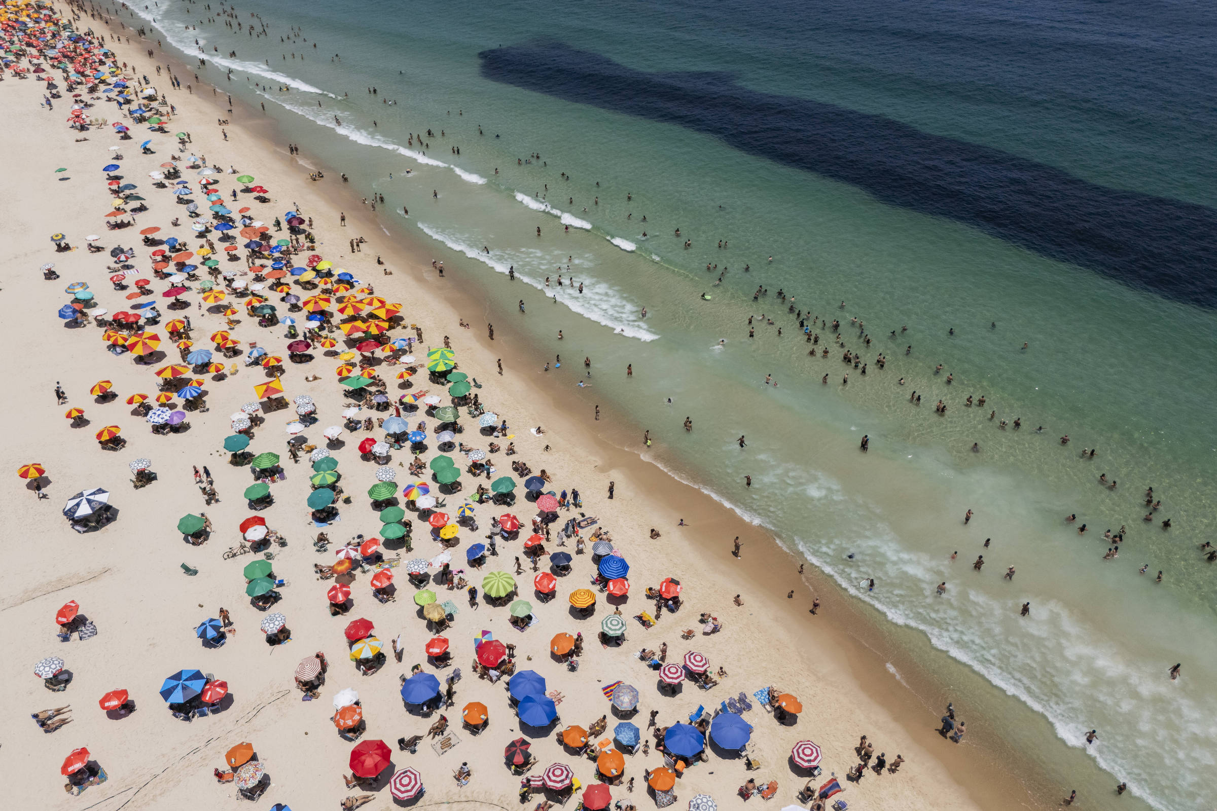 Vista aérea mostra praia cheia de guarda-sóis coloridos distribuídos na areia e muitas pessoas na água e na faixa de areia. O mar apresenta áreas claras e escuras, com ondas suaves chegando à praia.