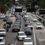 Rodízio de veículos em SP suspenso até sexta-feira 9 Vários veículos, incluindo carros e motos, formam congestionamento em avenida de pista dupla com faixa exclusiva para ônibus à direita. Árvores e construções cercam a via em dia nublado.