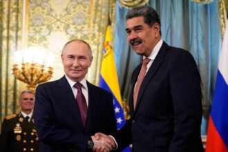 Vladimir Putin e Nicolás Maduro apertam as mãos em sala oficial com bandeiras da Venezuela e Rússia ao fundo. Militar observa ao lado.