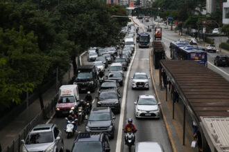 Veículos e motocicletas congestionados em duas faixas da via expressa durante o dia. Ônibus circulam na faixa exclusiva ao lado direito, com ponto de ônibus visível. Árvores e edifícios cercam a via.