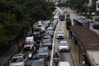 Veículos e motocicletas congestionados em duas faixas da via expressa durante o dia. Ônibus circulam na faixa exclusiva ao lado direito, com ponto de ônibus visível. Árvores e edifícios cercam a via.