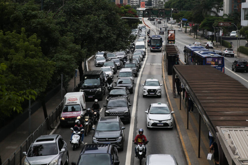 Veículos e motocicletas congestionados em duas faixas da via expressa durante o dia. Ônibus circulam na faixa exclusiva ao lado direito, com ponto de ônibus visível. Árvores e edifícios cercam a via.