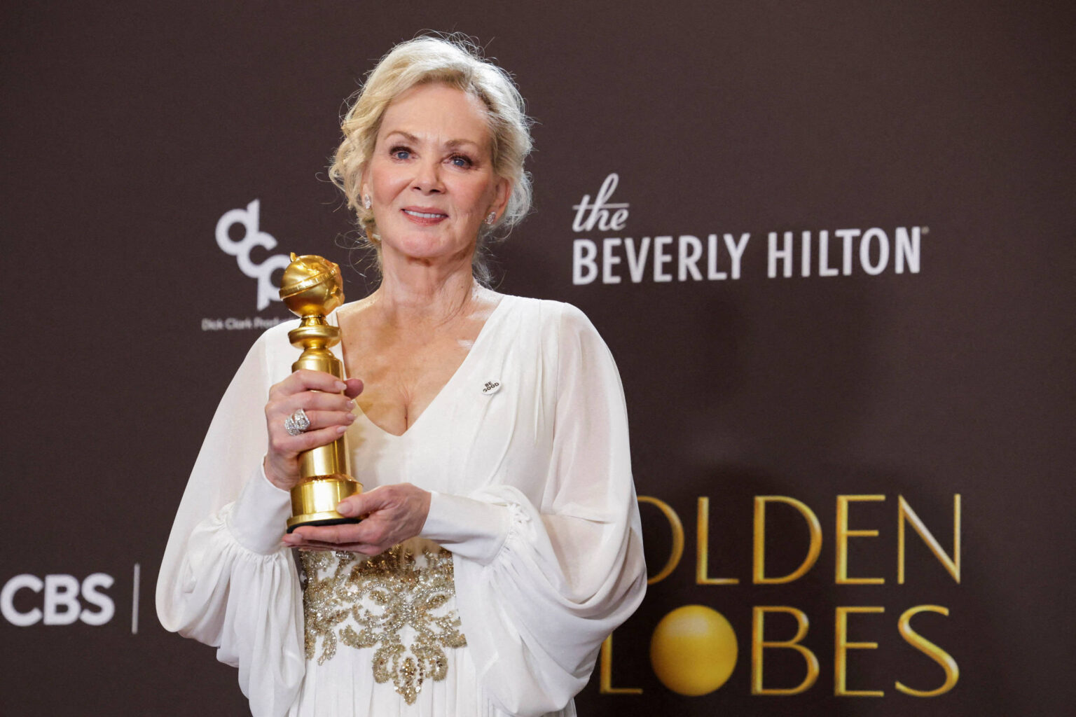 Mulher loira em vestido branco com detalhes dourados segura estatueta do Globo de Ouro em frente a fundo com logos do Beverly Hilton e Golden Globes.