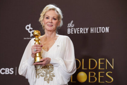 Mulher loira em vestido branco com detalhes dourados segura estatueta do Globo de Ouro em frente a fundo com logos do Beverly Hilton e Golden Globes.