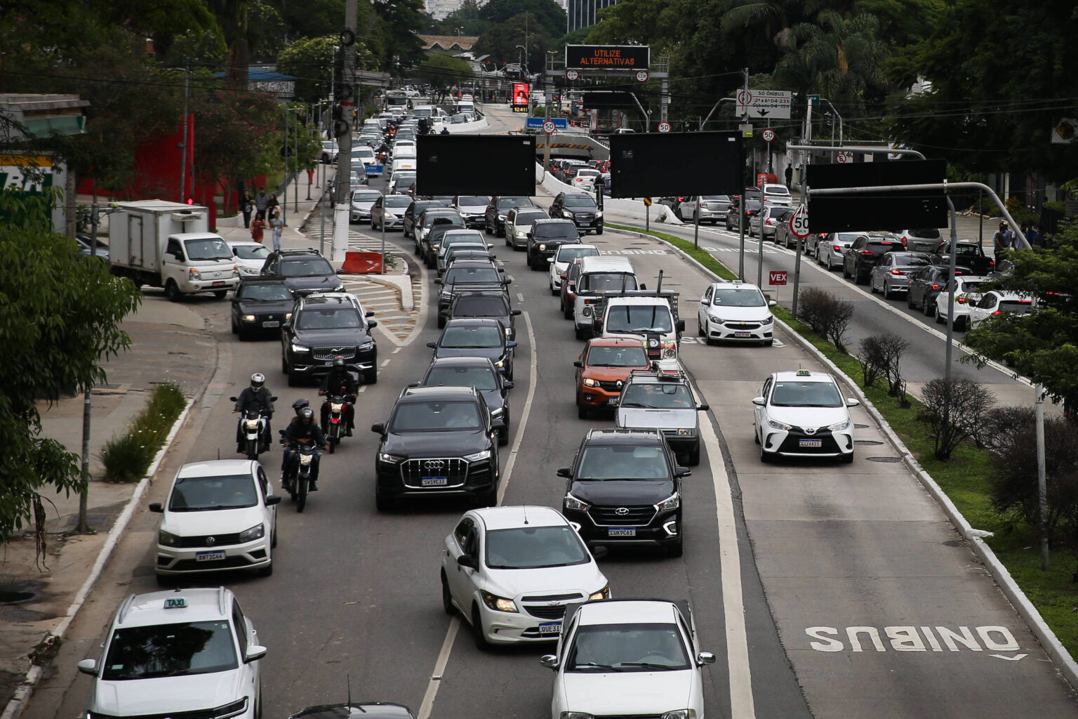 Rodízio de veículos em SP é retomado nesta segunda-feira Vários veículos, incluindo carros e motos, formam congestionamento em avenida de pista dupla com faixa exclusiva para ônibus à direita. Árvores e construções cercam a via em dia nublado.