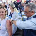 Homem aplica vacina no braço de mulher sorridente em ambiente com várias pessoas, incluindo profissionais de saúde com jalecos brancos e público ao fundo.