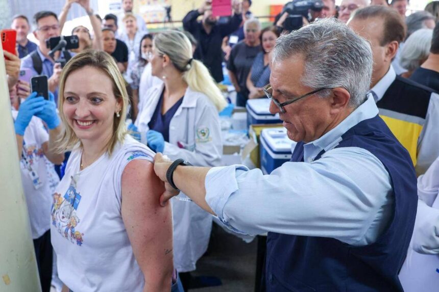 Homem aplica vacina no braço de mulher sorridente em ambiente com várias pessoas, incluindo profissionais de saúde com jalecos brancos e público ao fundo.