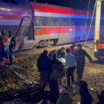 Trem com pintura vermelha, branca e azul parado nos trilhos à noite. Pessoas e equipes de resgate estão próximas ao trem e a uma ambulância com luzes ligadas.