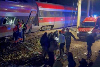 Trem com pintura vermelha, branca e azul parado nos trilhos à noite. Pessoas e equipes de resgate estão próximas ao trem e a uma ambulância com luzes ligadas.