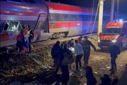 Trem com pintura vermelha, branca e azul parado nos trilhos à noite. Pessoas e equipes de resgate estão próximas ao trem e a uma ambulância com luzes ligadas.