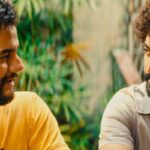 Dois homens sentados frente a frente em ambiente externo com plantas ao fundo. Um veste camiseta amarela e o outro camisa cinza clara, ambos com barba e cabelos encaracolados, olhando um para o outro.