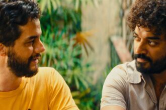 Dois homens sentados frente a frente em ambiente externo com plantas ao fundo. Um veste camiseta amarela e o outro camisa cinza clara, ambos com barba e cabelos encaracolados, olhando um para o outro.
