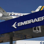 Embraer registra recorde na carteira de encomendas em 2025 Avião de asa alta com pintura branca e detalhes em azul e amarelo voa sobre estrutura azul com logo e nome da Embraer em branco. Céu azul ao fundo.