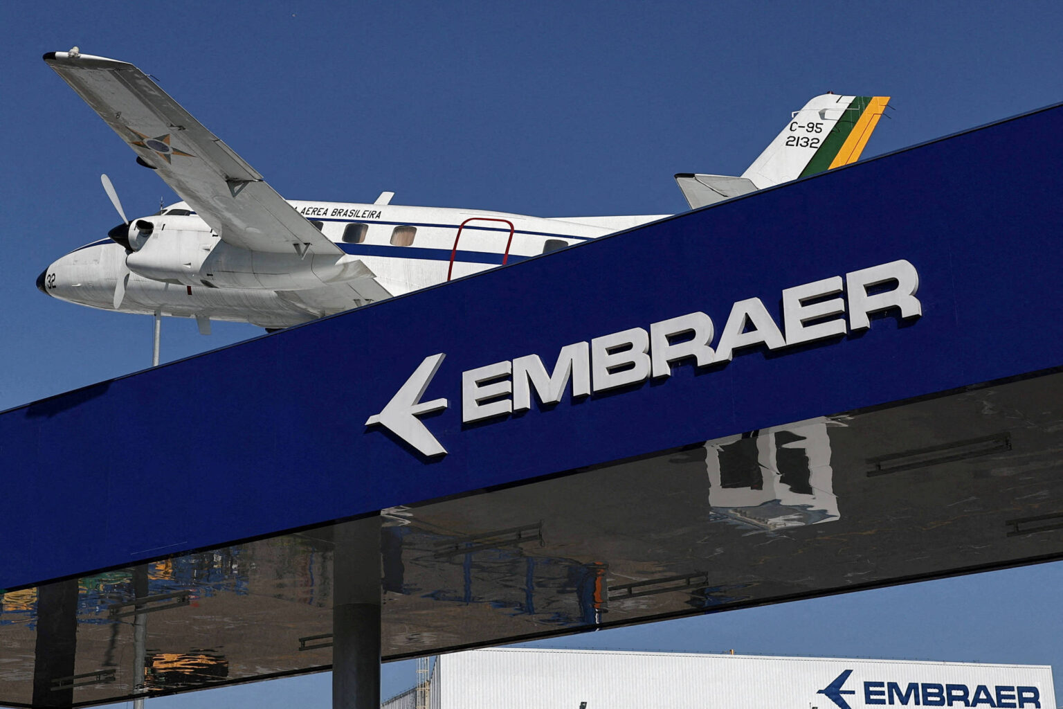Embraer registra recorde na carteira de encomendas em 2025 Avião de asa alta com pintura branca e detalhes em azul e amarelo voa sobre estrutura azul com logo e nome da Embraer em branco. Céu azul ao fundo.