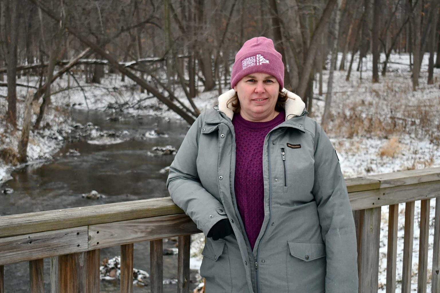 Exploração tranquila: junte-se a mim por um ano de aventuras A person stands at a bridge over a creek, wearing a grey coat and pink hat.