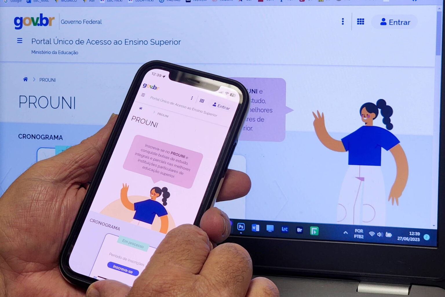 Prouni 2026: resultados da 1ª chamada são divulgados hoje Mãos seguram celular mostrando a página do PROUNI com ilustração de mulher de camiseta azul e calça branca. Ao fundo, laptop exibe a mesma página do PROUNI no Portal do Governo Federal.